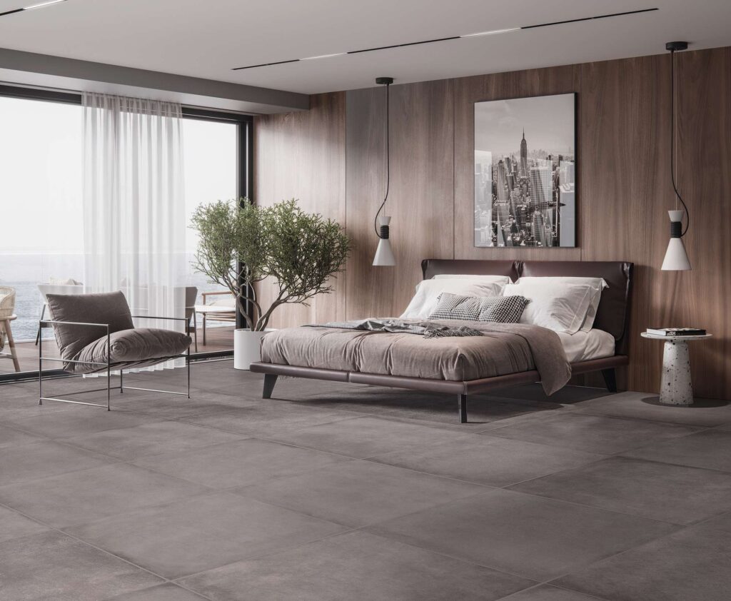 La collezione XO CEMENT nel formato 60X60 definisce il carattere di questa camera da letto di design, grazie a un pavimento in gres porcellanato grigio scuro effetto cemento con posa regolare a giunto unito. L'ambiente, dall'atmosfera calda e contemporanea, presenta una parete retrostante rivestita in legno pregiato su cui spicca un quadro metropolitano in bianco e nero. Un letto matrimoniale con testiera in pelle scura, un'elegante poltrona minimalista e un'ampia vetrata scorrevole che si affaccia su un terrazzo vista mare completano la stanza, inondandola di luce naturale.
