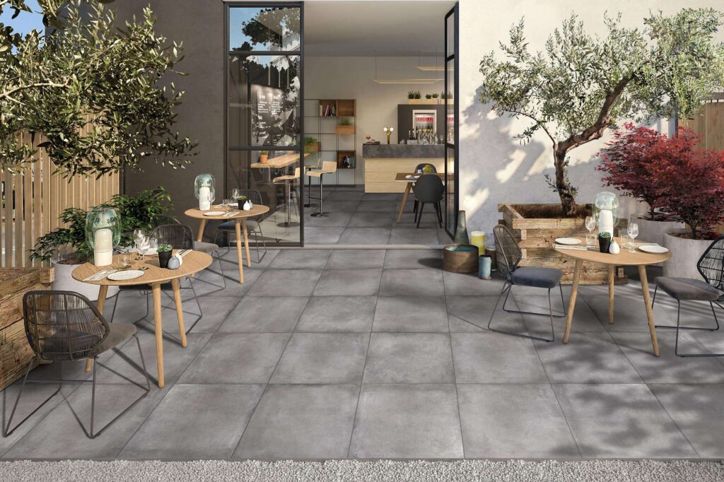 La collezione XO CEMENT, nel formato 60x60, valorizza la pavimentazione esterna di un elegante bistrot con le sue piastrelle in gres porcellanato effetto cemento grigio. L'immagine mostra un accogliente dehors soleggiato, arredato con tavolini rotondi in legno, sedie di design e alberi di ulivo in grandi vasi. La posa regolare delle piastrelle crea una perfetta continuità visiva e cromatica tra la terrazza esterna e l'interno del locale, visibile attraverso un'ampia vetrata aperta che conferisce profondità e un senso di moderno minimalismo all'ambiente.