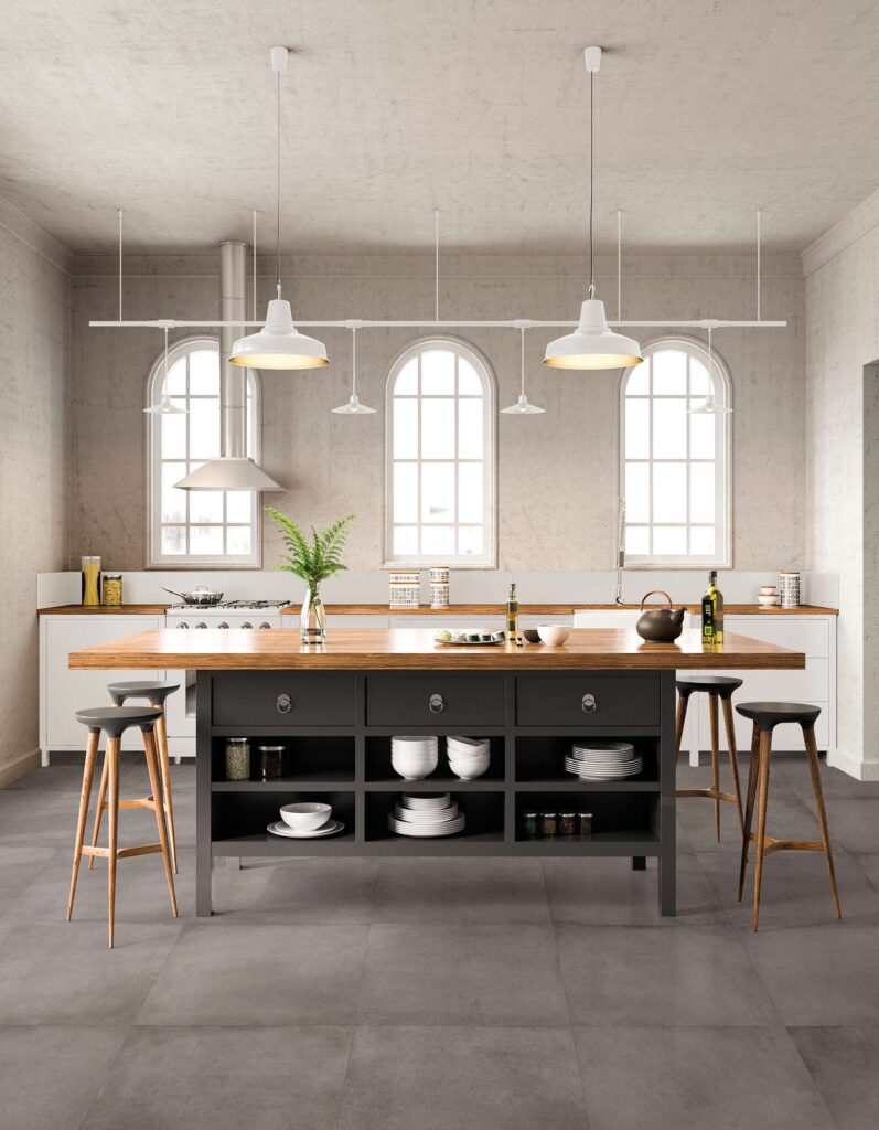La collezione XO CEMENT, nel formato 60x60 cm con effetto cemento grigio scuro, valorizza il pavimento di una spaziosa e luminosa cucina di design. L'ambiente presenta una grande isola centrale con piano in legno e base scura attrezzata, circondata da sgabelli alti. Sullo sfondo, ampie finestre ad arco inondano di luce naturale la stanza, esaltando la posa regolare delle piastrelle e la texture materica della pavimentazione, per un'atmosfera contemporanea e accogliente.
