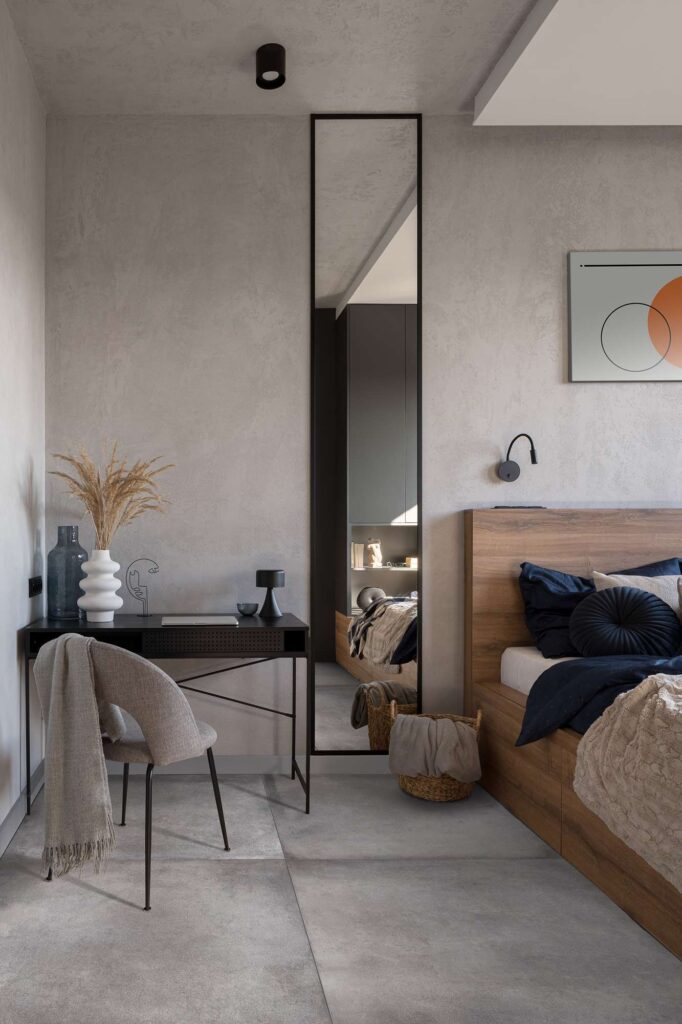 La collezione di gres porcellanato XO Cement, nel formato 80x80 cm con finitura grigia effetto cemento, riveste il pavimento di una raffinata camera da letto dal design contemporaneo. La posa lineare delle grandi lastre valorizza la texture materica e minimalista dell'ambiente, caratterizzato da un letto in legno caldo con tessuti blu scuro, un elegante specchio verticale a tutta altezza e un angolo scrittoio nero con sedia coordinata e dettagli decorativi. Un'atmosfera accogliente, armoniosa e di alto design.