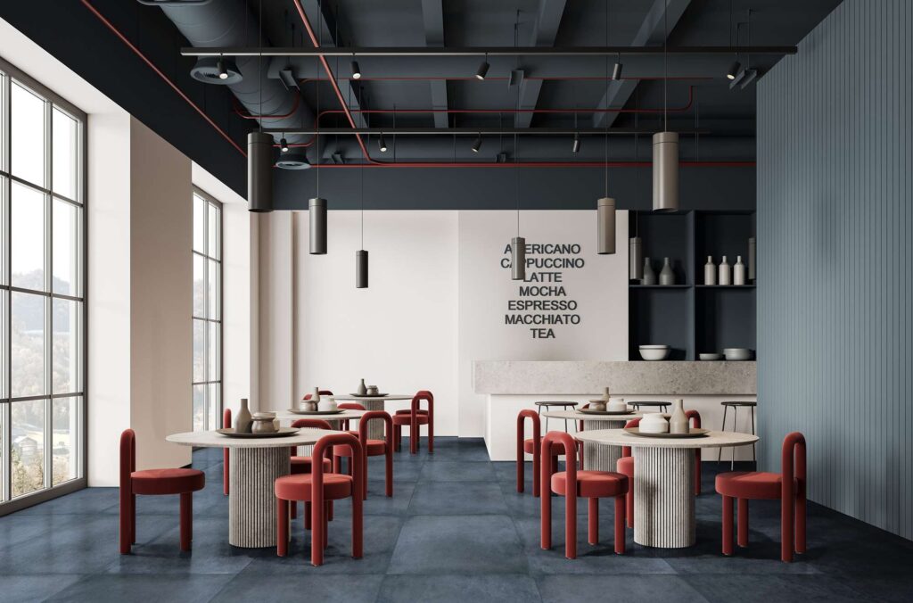 La collezione XO Midnight nel grande formato 120x120 cm definisce la pavimentazione di un ristorante moderno dal design ricercato. Le piastrelle in gres porcellanato nella scura e intensa tonalità blu notte sono posate con fuga minima, creando una superficie continua che contrasta elegantemente con le sedute di design in velluto rosso mattone e i tavoli rotondi chiari. L'ambiente, luminoso grazie alle ampie vetrate laterali, è caratterizzato da un soffitto industriale scuro con condutture a vista e luci a sospensione cilindriche, per un'atmosfera sofisticata e contemporanea.