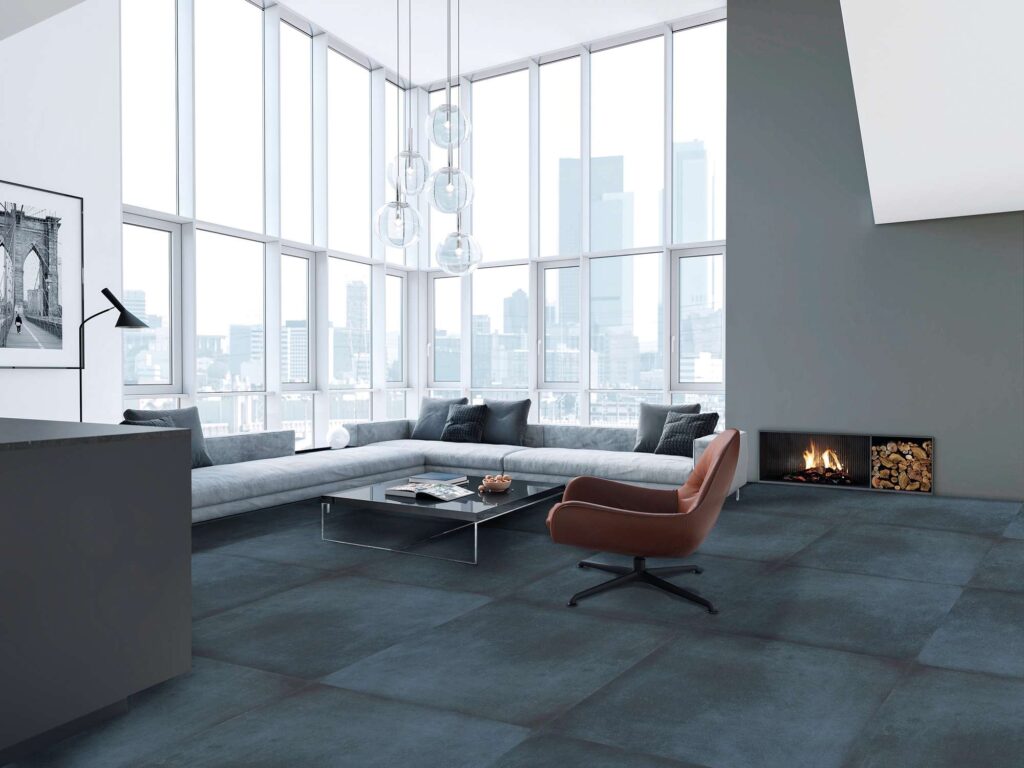 La collezione XO in variante Midnight nel formato 60x60 riveste il pavimento di un prestigioso e luminoso ambiente living. Le piastrelle in gres porcellanato scuro dalle sfumature blu notte, posate in modo regolare, donano profondità e contrastano elegantemente con la luce naturale che inonda la stanza dalle grandi vetrate panoramiche con vista sullo skyline urbano. L'arredamento contemporaneo d'eccellenza è composto da un ampio divano angolare grigio chiaro, una poltrona di design in pelle marrone, un tavolino basso nero e un moderno camino a parete integrato, che insieme creano un'atmosfera lussuosa e accogliente.