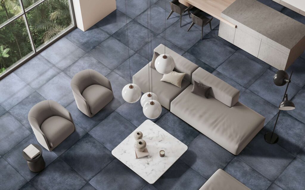 La collezione XO Midnight 80X80 veste il pavimento di un elegante e luminoso ambiente living con piastrelle in gres porcellanato dalle intense sfumature blu scuro ed effetto materico. La vista dall'alto mostra un soggiorno di design contemporaneo caratterizzato da ampie vetrate affacciate sulla natura, arredato con un divano componibile e poltrone coordinate nei toni del beige, un tavolino quadrato in marmo bianco e lampade a sospensione sferiche. La posa regolare dei grandi formati quadrati esalta la continuità visiva e la raffinatezza dello spazio.