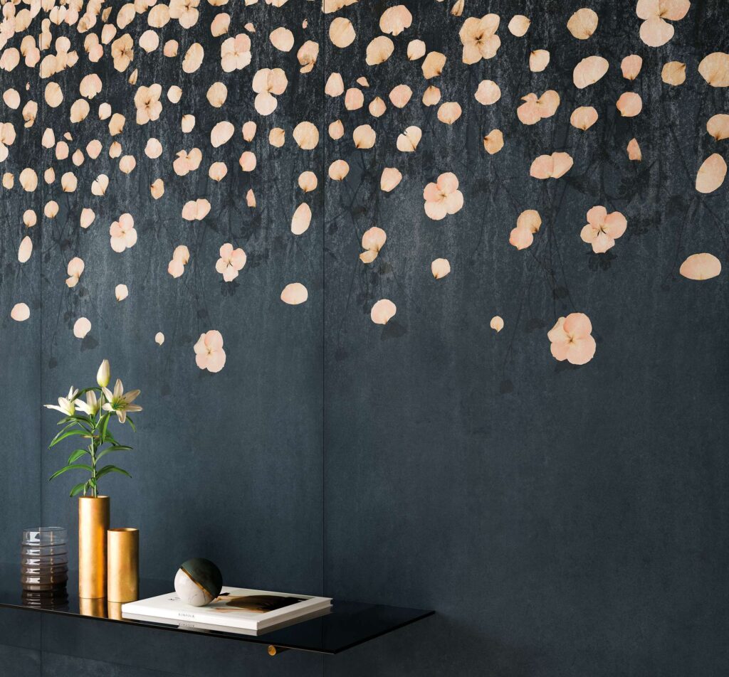 La collezione XO Midnight nella variante decorativa Dec. Cascade, nel formato 60x120 cm, riveste la parete con un elegante gres porcellanato scuro, color blu notte e antracite. La superficie è impreziosita da una poetica cascata di petali e piccoli fiori nei toni delicati del rosa cipria e del beige che scendono dall'alto. In primo piano, un allestimento raffinato mostra una mensola nera di design con vasi cilindrici dorati che ospitano gigli bianchi freschi, una sfera decorativa e un libro d'arte, creando un'atmosfera intima, lussuosa e contemporanea.