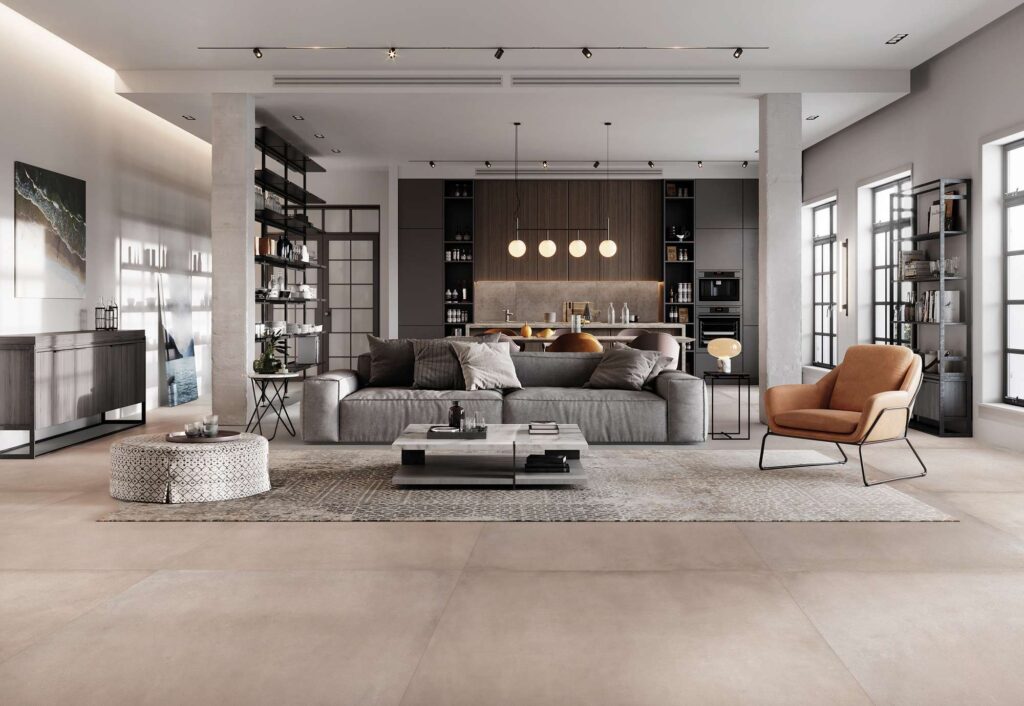 La collezione di piastrelle in gres porcellanato XO Poudre, nel grande formato 120x120 cm, riveste il pavimento di un elegante e luminoso open space adibito a living. L'ambiente moderno e sofisticato presenta un ampio divano grigio posizionato su un tappeto decorato, un tavolino centrale in marmo e una poltrona di design color cognac. Grandi colonne in cemento a vista incorniciano lo spazio, che si apre sullo sfondo verso una cucina contemporanea e ampie vetrate in stile industriale. La luce naturale esalta la superficie calda e uniforme della pavimentazione ceramica color beige cipria (Poudre).