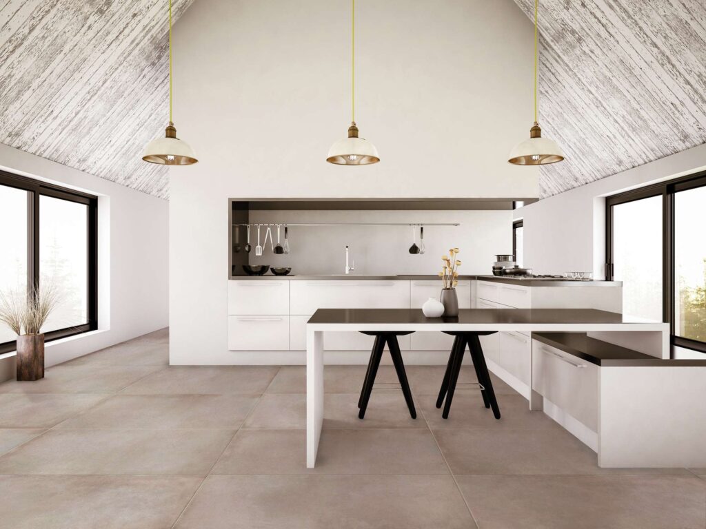 La collezione XO Poudre nel formato 60x120 cm valorizza il pavimento di una cucina moderna e minimalista inondata di luce naturale. Le piastrelle in gres porcellanato, caratterizzate dalla delicata tonalità grigio-beige 'Poudre' effetto cemento, sono posate in modo regolare per enfatizzare l'ampiezza dello spazio. L'ambiente presenta un'isola centrale con piano nero e sgabelli coordinati, arredi bianchi essenziali, un soffitto spiovente in legno decapato e tre eleganti lampade a sospensione, per un’atmosfera raffinata e contemporanea.