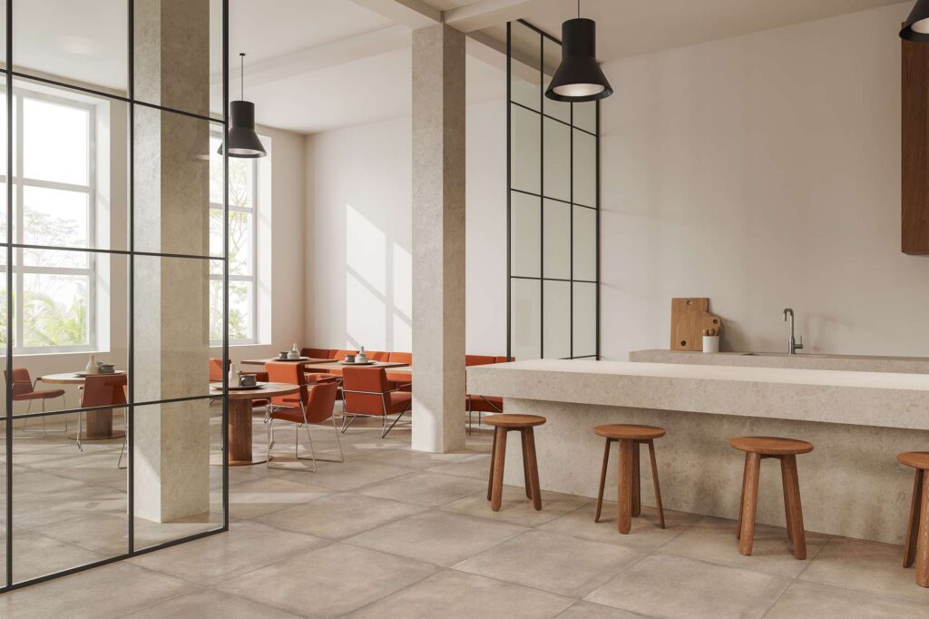 La collezione di piastrelle XO POUDRE nel formato 80x80 cm veste il pavimento di un moderno e raffinato bar dal design minimalista. La posa lineare e regolare mette in risalto la calda tonalità cipria e la texture effetto pietra della superficie ceramica. L'ambiente, luminoso e accogliente, è caratterizzato da un imponente bancone materico completato da sgabelli in legno, eleganti divisori in vetro con profili metallici neri e una zona tavoli con sedute color terracotta.