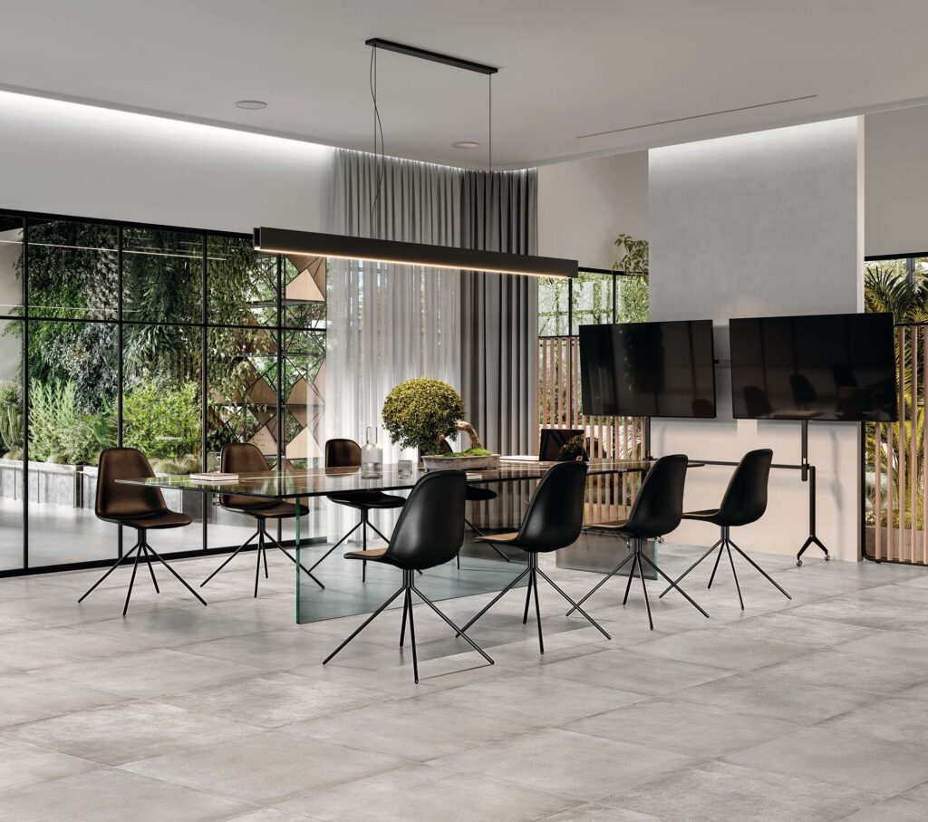La collezione di piastrelle XO Smoke in formato 60x60 cm riveste con eleganza il pavimento di un moderno e luminoso ambiente living e sala riunioni. La pavimentazione in gres porcellanato grigio chiaro effetto cemento, posata in modo regolare, fa da base a un grande tavolo di design con piano in vetro e sedie nere minimaliste. Sullo sfondo, ampie vetrate a tutta altezza si affacciano su un rigoglioso giardino verticale, infondendo un senso di naturalezza e raffinatezza all'intero spazio contemporaneo.