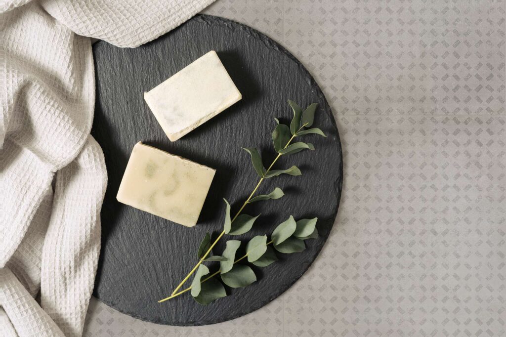 La collezione XO nella variante Smoke Grid nel formato 60x60 cm si rivela in un elegante e minimalista piano ravvicinato. La superficie della piastrella grigia, arricchita da una texture geometrica discreta, fa da sfondo a una raffinata composizione wellness in stile flat lay, con due saponette artigianali adagiate su un vassoio circolare di ardesia scura, un rametto di eucalipto fresco e un soffice telo a nido d'ape chiaro.