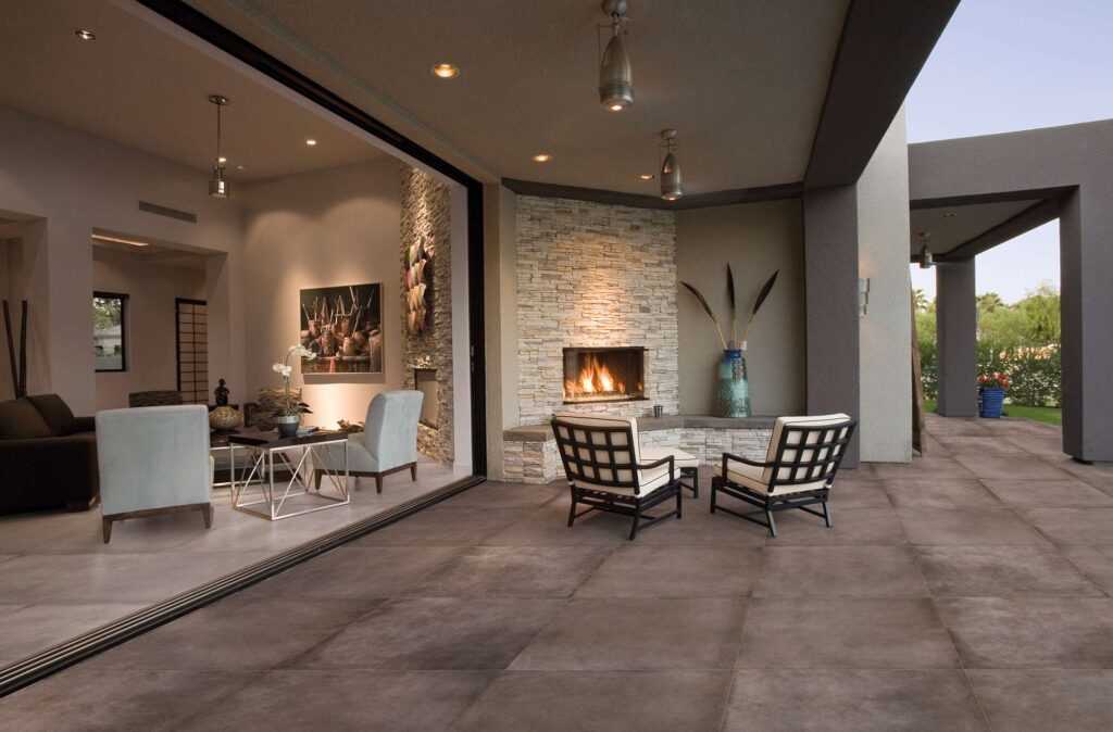La collezione XO, nella calda e profonda tonalità Tobacco in formato 60x60, riveste con eleganza il pavimento di un moderno patio esterno coperto. La posa regolare delle grandi piastrelle esalta lo spazio relax all'aperto, arredato con poltrone di design e un suggestivo camino ad angolo in pietra con fiamma accesa. La grande vetrata scorrevole aperta mostra la perfetta continuità visiva tra l'esterno e il lussuoso soggiorno interno, creando un'atmosfera calda, accogliente e di grande prestigio.