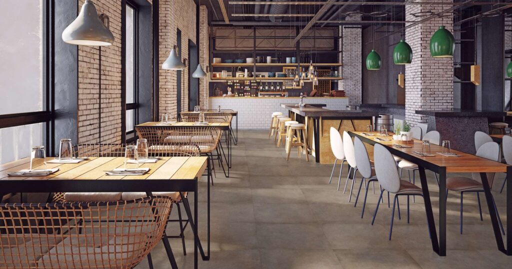 La collezione di pavimenti in gres porcellanato XO TOBACCO, nel formato 60x60 cm, valorizza l'ampia sala di un moderno ristorante dal raffinato design industrial-chic. Le piastrelle nella calda tonalità tobacco, posate con uno schema regolare e ortogonale, si estendono su tutta la superficie donando continuità visiva. L'ambiente è caratterizzato da tavoli in legno naturale accostati a sedie di design, pareti con mattoncini bianchi a vista, grandi vetrate che diffondono luce naturale e un accogliente bancone bar sullo sfondo con sgabelli in legno.