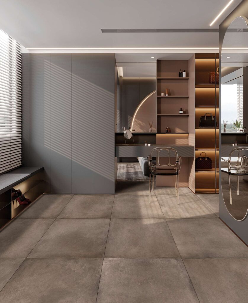 La collezione XO TOBACCO 80X80 presenta un pavimento in gres porcellanato nel formato 80x80 cm nella calda e avvolgente tonalità Tobacco. Le piastrelle, posate con fuga minima in un ambiente moderno e lussuoso dedicato alla zona vanity e cabina armadio, mostrano una texture materica e naturale. La stanza è arredata con eleganza minimalista, caratterizzata da una postazione trucco con sedia trasparente di design, armadiature grigie a tutta altezza e scaffali retroilluminati che espongono accessori preziosi. La luce naturale, filtrata da una tenda veneziana, crea un suggestivo gioco di ombre geometriche che valorizza la superficie della ceramica, conferendo un'atmosfera calda, sofisticata e accogliente.