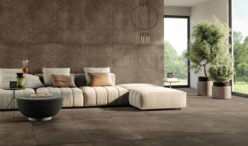 La collezione XO TOBACCO con decoro DEC. ARABESQUE nel formato 60x120 riveste con eleganza questo moderno e luminoso ambiente living. La pavimentazione in gres porcellanato nei toni caldi del marrone tabacco si coordina con la parete di fondo, impreziosita da piastrelle decorate con un raffinato motivo damascato arabesco tono su tono. L'ambiente, dall'atmosfera calda e accogliente, è arredato con un ampio divano componibile color panna, tavolini bassi di design e piante verdi in vaso, il tutto illuminato dalla luce naturale che filtra dalle ampie vetrate affacciate sul giardino.