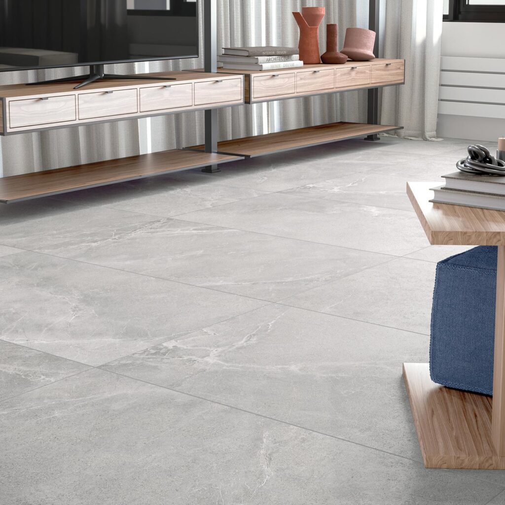 La collezione BLEND, proposta nella sfumatura grigio cenere Ash nel formato 60x120 cm, veste il pavimento di un moderno soggiorno (living) con raffinato gres porcellanato effetto pietra. L'inquadratura in primo piano mette in risalto la posa delle grandi lastre rettangolari caratterizzate da delicate venature naturali, inserite in un ambiente luminoso con un mobile TV sospeso in legno e metallo, tende chiare e complementi d'arredo contemporanei.