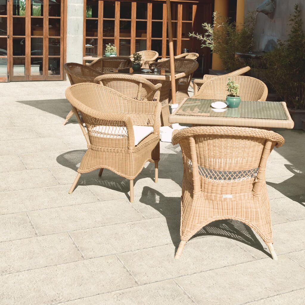 La collezione Bonnieux Beige, nel formato 30x60 per esterni, caratterizza la pavimentazione in gres porcellanato effetto pietra di un raffinato patio soleggiato. Le piastrelle, posate con schema sfalsato, mostrano una texture materica e naturale color beige che si integra armoniosamente con l'arredo outdoor composto da eleganti poltroncine e tavoli in rattan, piante decorative e ampie vetrate sullo sfondo, creando un'atmosfera accogliente e di classe