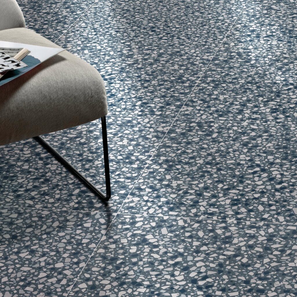 La collezione D Lash presenta il gres porcellanato Mineral Blue nel formato 60x60, caratterizzato da un raffinato effetto terrazzo alla veneziana con graniglia bianca, azzurra e grigia su un fondo blu minerale. L'immagine mostra un dettaglio della pavimentazione posata con precisione, accostata a una moderna sedia imbottita in tessuto grigio chiaro con gambe in metallo nero, creando un'atmosfera contemporanea e luminosa grazie alla luce naturale che si riflette sulla superficie ceramica.