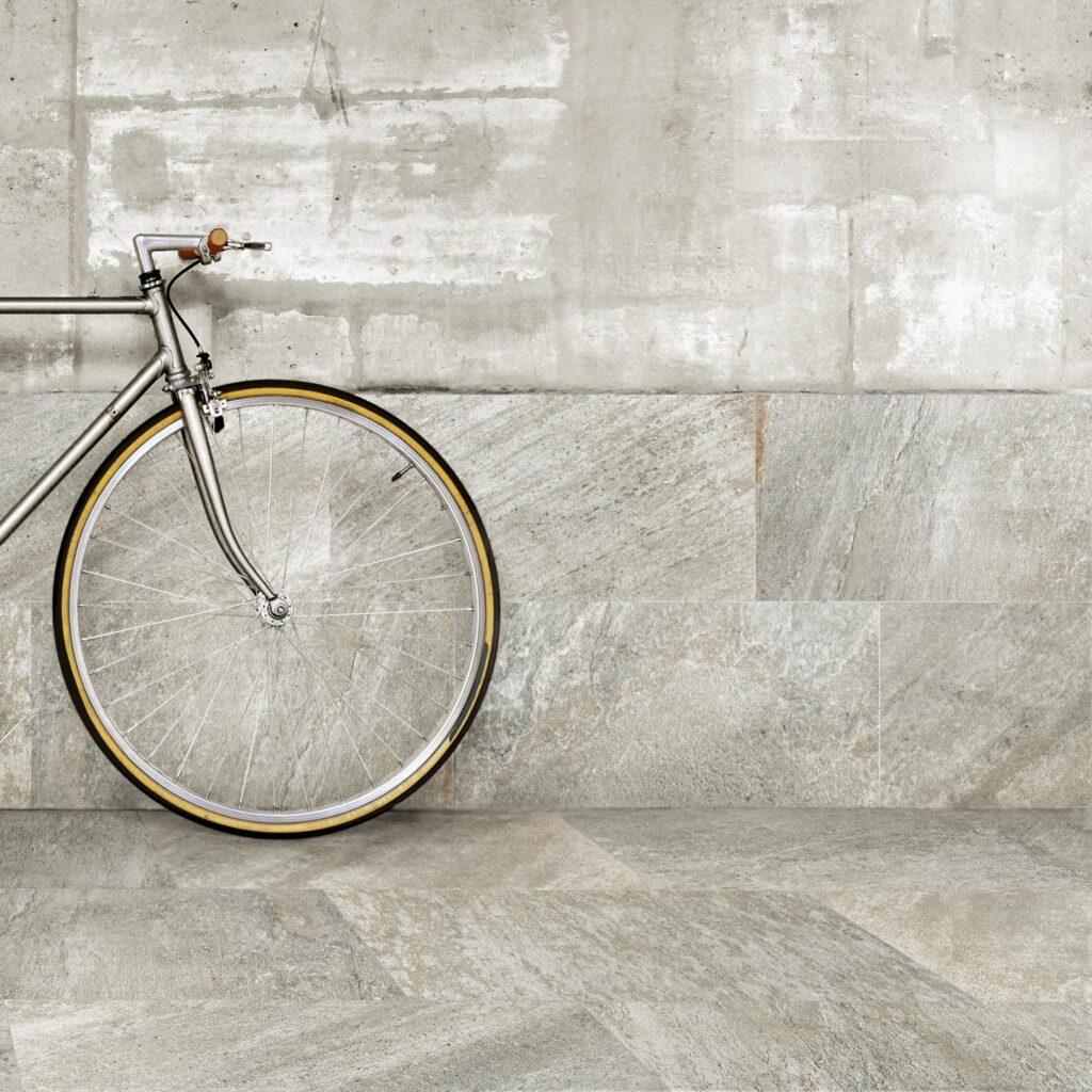 La collezione Discovery Colonial nel formato 30x60 cm valorizza questo spazio d'ispirazione urban-chic con piastrelle in gres porcellanato effetto pietra dalle ricche sfumature grigie e naturali. Nell'immagine, una bicicletta vintage dal telaio metallico è appoggiata a una parete parzialmente rivestita con posa orizzontale, creando una continuità armonica con il pavimento coordinato e mettendo in risalto la texture materica e antiscivolo della superficie.