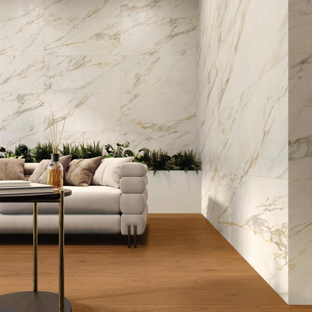 La collezione Amb Aurea definisce un elegante spazio living dove il gres porcellanato effetto legno Barrique Cherry nel formato 20X120 a pavimento incontra il raffinato rivestimento a parete effetto marmo Elysion Calacatta nel formato 60x120. L'ambiente, luminoso e moderno, mostra un angolo salotto con un divano grigio chiaro arricchito da soffici cuscini, una fioriera con piante verdi sullo sfondo e un tavolino di design in primo piano. Le venature calde e dorate della parete effetto marmo si coordinano armoniosamente con le tonalità calde del pavimento in legno, creando un'atmosfera accogliente e di grande prestigio.