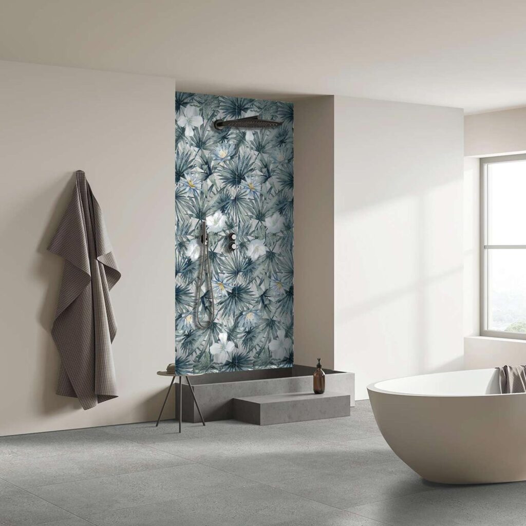 La collezione I Decors veste un elegante e moderno ambiente bagno, dove spicca il contrasto armonioso tra natura e minimalismo. All'interno della doccia, la parete di fondo è rivestita con il decoro floreale Maui Sea nel formato 60x120 cm, caratterizzato da un raffinato motivo di foglie tropicali e fiori bianchi su fondo blu-grigio. A pavimento, la posa delle piastrelle Mon Smoke nel formato 60x120 cm offre una superficie materica grigia che dona sobrietà e continuità visiva all'ambiente. Lo spazio, ampio e inondato di luce naturale grazie a una grande finestra, ospita una vasca freestanding bianca di design, evocando un'atmosfera di puro benessere e raffinata spa domestica.