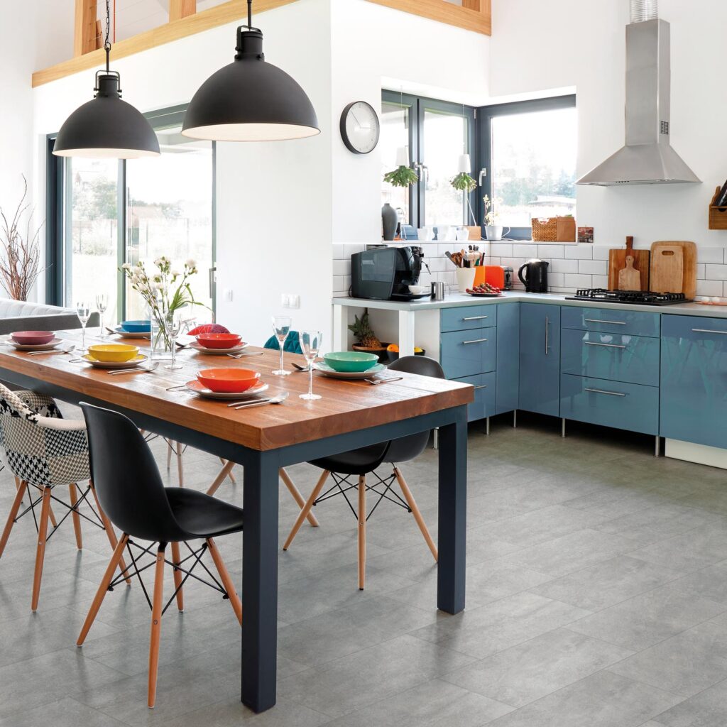 Pavimento in gres porcellanato della collezione Jelling Silver nel formato 30x60 cm, caratterizzato da eleganti piastrelle grigie effetto pietra con posa a correre. La superficie valorizza una spaziosa e luminosa cucina open space con zona pranzo, arredata con un grande tavolo in legno massiccio, sedie di design contemporaneo e dettagli moderni, che si apre su un'accogliente area living con ampie vetrate sul giardino.
