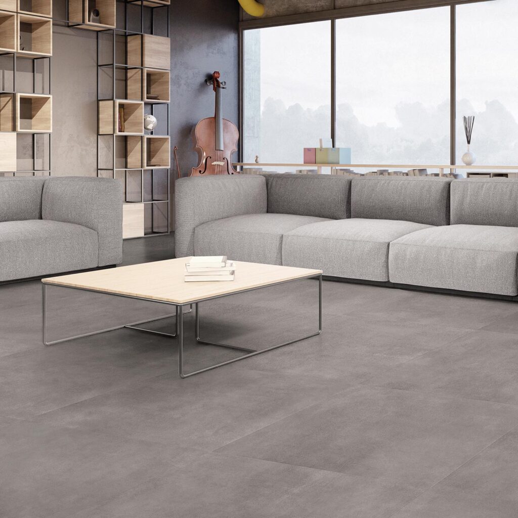 La collezione Materia nella tonalità Grafite, nel formato 60x60 cm, riveste il pavimento di un raffinato e luminoso ambiente living dal design contemporaneo. Le piastrelle in gres porcellanato grigio scuro, caratterizzate da una texture materica e posate con precisione, fanno da cornice a un arredamento moderno e minimalista: un ampio divano modulare grigio chiaro, un tavolino basso con top in legno e un'elegante libreria a giorno in metallo e legno. Sullo sfondo, una grande vetrata a tutta altezza inonda lo spazio di luce naturale, valorizzando l'atmosfera sobria e sofisticata della stanza.