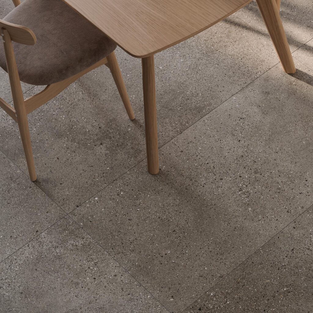 La collezione MON GREIGE nel formato 60x120 cm veste il pavimento di una raffinata zona pranzo adiacente alla cucina. Le grandi lastre in gres porcellanato effetto pietra in tonalità greige, caratterizzate da una texture materica finemente puntinata, valorizzano l'ambiente accogliendo un tavolo in legno chiaro e sedie coordinate dal design contemporaneo. La luce naturale che filtra trasversalmente esalta i dettagli della posa e la superficie elegante e naturale delle piastrelle.