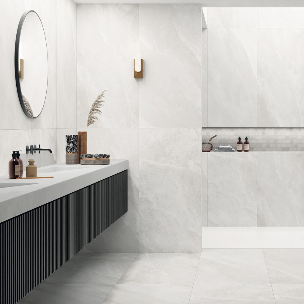 La collezione OCEAN BLACK PEARL veste questo elegante bagno moderno con grandi piastrelle in formato 60x120 cm dall'effetto pietra grigio chiaro e luminoso, posate sia a pavimento che a parete. Nella nicchia della doccia walk-in è inserito un raffinato dettaglio decorativo realizzato con il mosaico in formato 5x5 cm. L'arredamento minimalista, composto da un lungo mobile lavabo sospeso con finitura rigata scura, rubinetteria nera a contrasto e un pouf di design, esalta l'atmosfera luminosa, lussuosa e rilassante dell'ambiente.