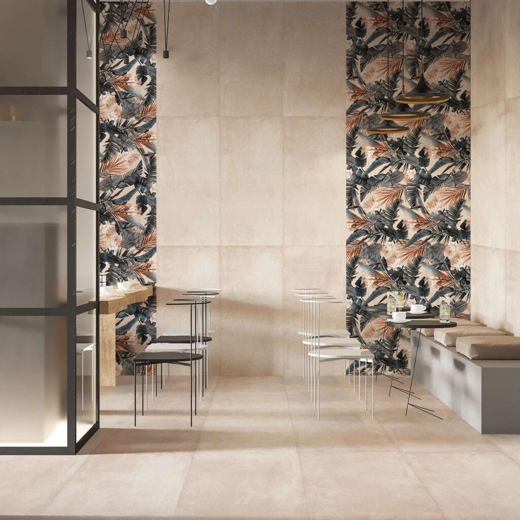 La collezione di piastrelle in gres porcellanato PROTON BEIGE, nel formato 60x120 cm, riveste con eleganza il pavimento e le pareti di questo moderno ristorante. La texture sobria e naturale nei toni del beige si sposa armoniosamente con le fasce decorative verticali a parete DEC.TEXDECO WARM, caratterizzate da un raffinato motivo botanico a fogliame nei toni caldi e naturali. L'ambiente, dall'atmosfera sofisticata e accogliente, è completato da arredi minimalisti come tavoli rotondi neri, sedie di design e una moderna panca con cuscini, valorizzati da una calda illuminazione sospesa.