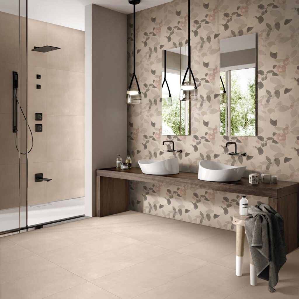 La collezione QUINTESSENCE in formato 60x120 cm nella sfumatura Beige riveste con eleganza il pavimento e la parete interna della doccia in questo bagno moderno e raffinato. La parete principale è valorizzata da un rivestimento decorativo a tema botanico, caratterizzato da un delicato motivo di foglie nei toni del grigio, beige e rosa antico. L'ambiente è completato da una consolle sospesa in legno scuro con due lavabi da appoggio bianchi, specchi verticali, lampade a sospensione nere e una doccia con eleganti dettagli in nero opaco.