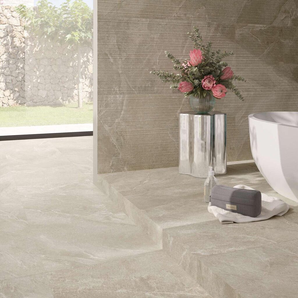 La collezione RESHAPE SAND 60x120 in gres porcellanato effetto pietra definisce lo stile di un bagno moderno e luminoso. La pavimentazione color sabbia a grandi lastre si sviluppa su un elegante piano rialzato, coordinandosi con il rivestimento a parete caratterizzato dalla raffinata texture a righe orizzontali "Lines". L'ambiente è completato da una vasca freestanding bianca di design, un tavolino metallico riflettente con un vaso di fiori rosa e un'ampia vetrata con vista su un giardino esterno che esalta la naturalezza e la luminosità del design.