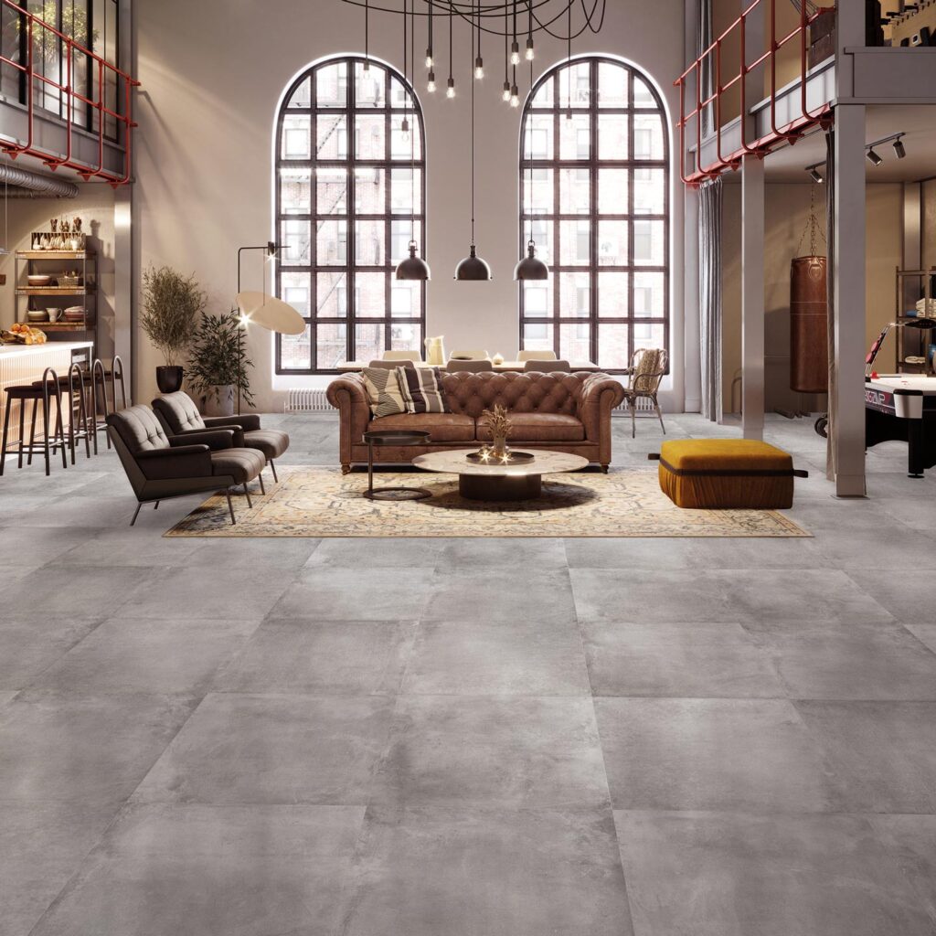 La collezione Unika, nella sfumatura grigia Smoke in formato 60x60 cm, definisce con eleganza il pavimento di un ampio e prestigioso soggiorno in stile loft industriale. Il gres porcellanato effetto pietra, posato in una griglia regolare e uniforme, riflette la luce naturale proveniente dalle imponenti finestre ad arco a doppia altezza. L'ambiente è valorizzato da arredi di classe, tra cui un classico divano Chesterfield in pelle marrone posizionato su un tappeto decorato, poltrone moderne e calde luci a sospensione, per un'atmosfera sofisticata, accogliente e dal design contemporaneo.
