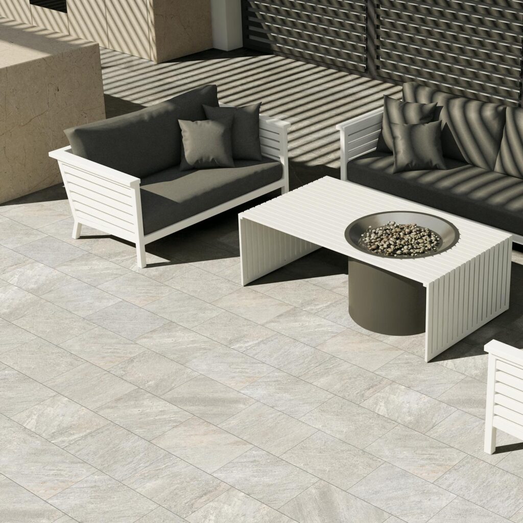 La collezione VENTIQUARANTA, nella raffinata tonalità Seattle Grey e nel formato 20x40, definisce lo stile di questo elegante spazio outdoor. La pavimentazione in gres porcellanato effetto pietra, caratterizzata da una posa a correre sfalsata, ospita un lussuoso salotto da giardino con divani e poltrone bianchi dai cuscini scuri, disposti attorno a un tavolino con braciere centrale. Sullo sfondo si scorge una cucina da esterno dal design minimale, mentre una luce solare radente proietta un suggestivo gioco di ombre geometriche sulla superficie, esaltando l'atmosfera sofisticata e accogliente del patio.