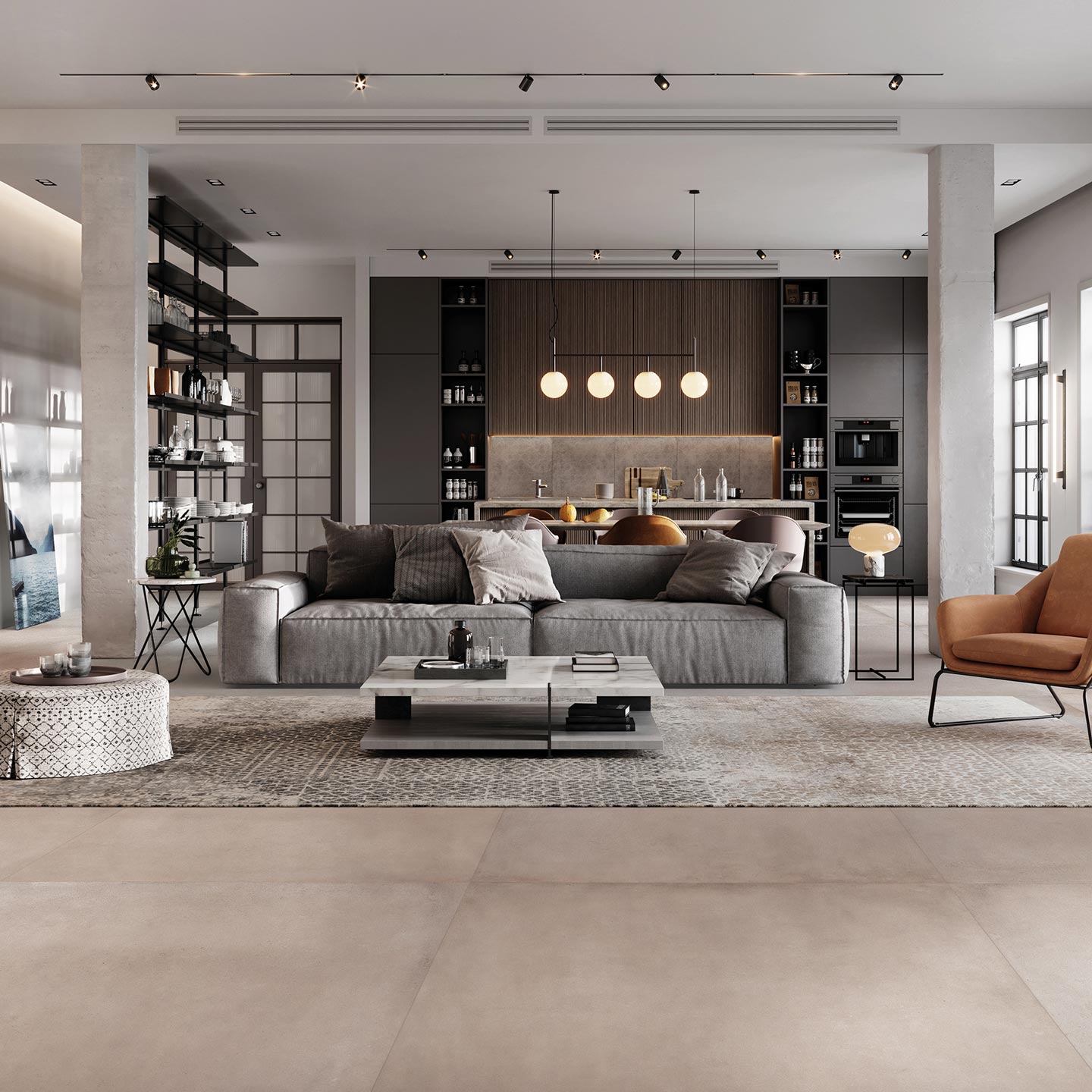 La collezione di piastrelle in gres porcellanato XO Poudre, nel grande formato 120x120 cm, riveste il pavimento di un elegante e luminoso open space adibito a living. L'ambiente moderno e sofisticato presenta un ampio divano grigio posizionato su un tappeto decorato, un tavolino centrale in marmo e una poltrona di design color cognac. Grandi colonne in cemento a vista incorniciano lo spazio, che si apre sullo sfondo verso una cucina contemporanea e ampie vetrate in stile industriale. La luce naturale esalta la superficie calda e uniforme della pavimentazione ceramica color beige cipria (Poudre).