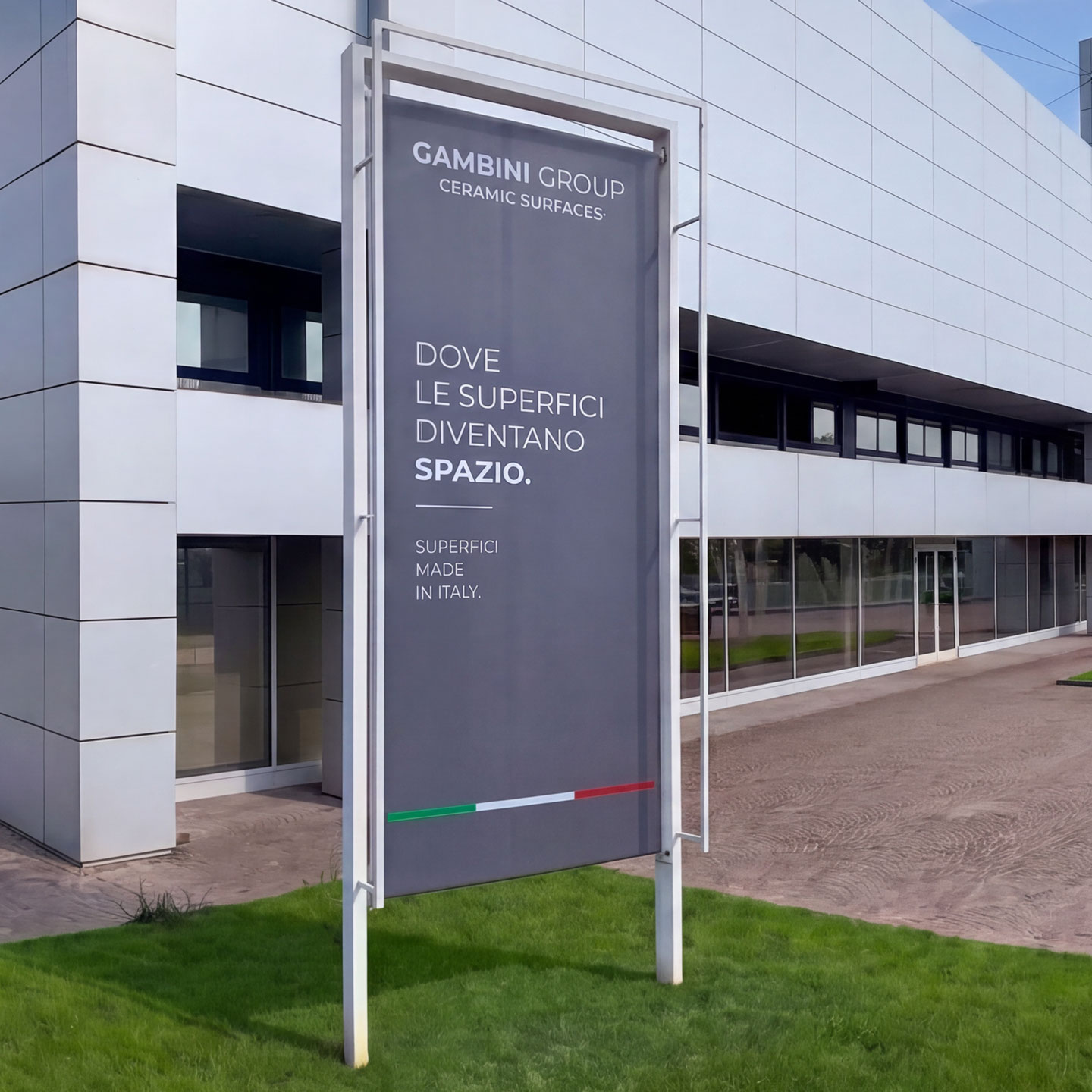 Totem istituzionale di Gambini Group posizionato davanti alla sede aziendale, con il messaggio "Dove le superfici diventano spazio", inserito in un contesto architettonico contemporaneo caratterizzato da facciate moderne, ampie vetrate e spazi esterni curati.